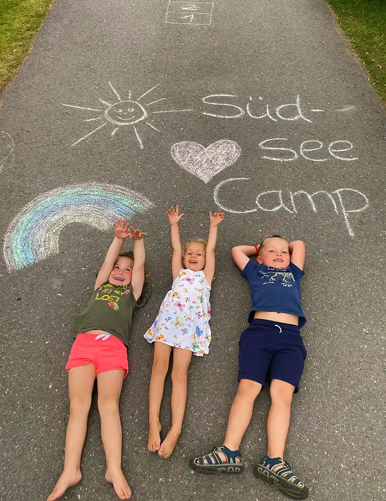Über uns Drei Kinder liegen barfuß lachend auf einem Asphaltweg, auf dem mit bunter Kreide eine Sonne, ein Herz, ein Regenbogen und der Schriftzug „Südsee-Camp“ gemalt wurden.