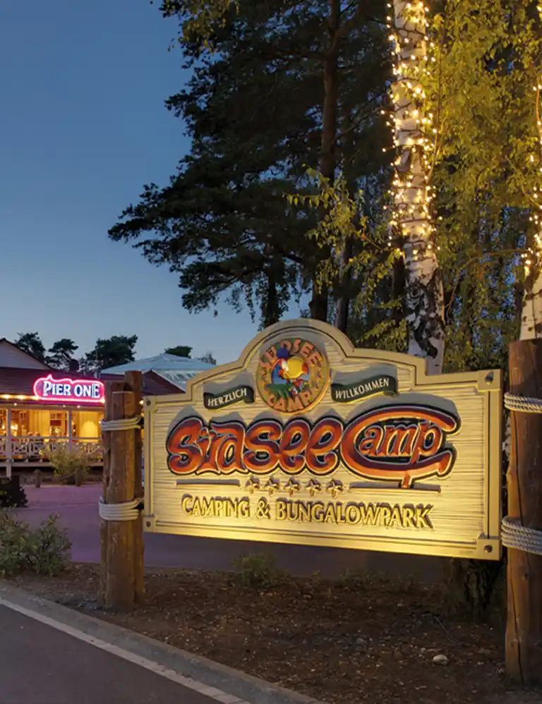 Lieblingsplatz finden Einfahrt zum Südsee-Camp bei Abenddämmerung mit beleuchtetem Willkommensschild und Lichterketten an den Bäumen.