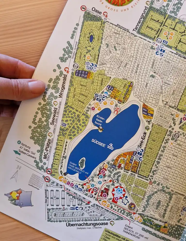 Übersicht verschaffen Übersichtskarte des Südsee-Camps mit eingezeichneten Bereichen wie dem Südsee-See, Campingplätzen, Sanitärgebäuden, Spielplätzen und thematisch benannten Arealen wie „Seewald“, „Wrogewald“ und „Oase“. Eine Hand hält die Karte am Rand fest.