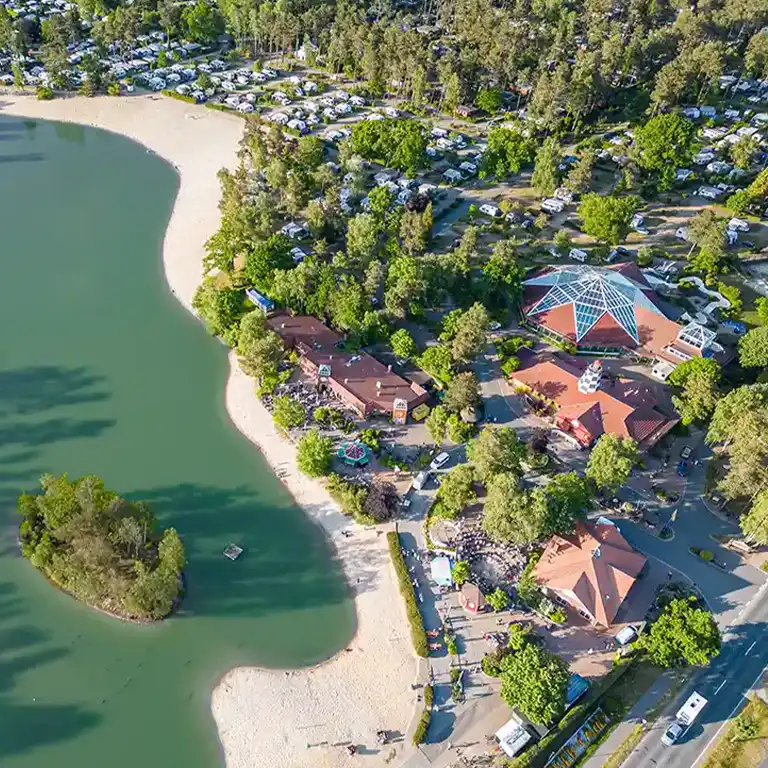 weitere Platzgebiete ansehen Luftaufnahme des Südsee-Camps mit Blick auf das zentrale Schwimmbad unter einem markanten Glasdach. Umgeben von Bäumen, Campingstellplätzen und einem großen Badesee mit Sandstrand. Die gepflegte Anlage bietet zahlreiche Freizeitmöglichkeiten inmitten der Natur.
