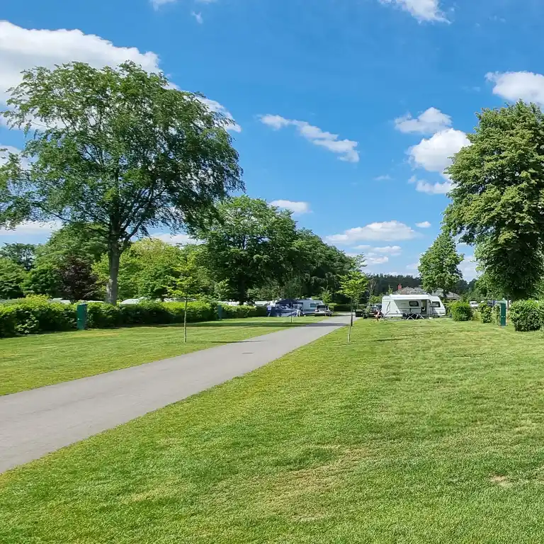Detailplan runterladen Großzügiger Campingplatz mit Wohnwagen, grüner Wiese und asphaltierter Straße unter blauem Himmel