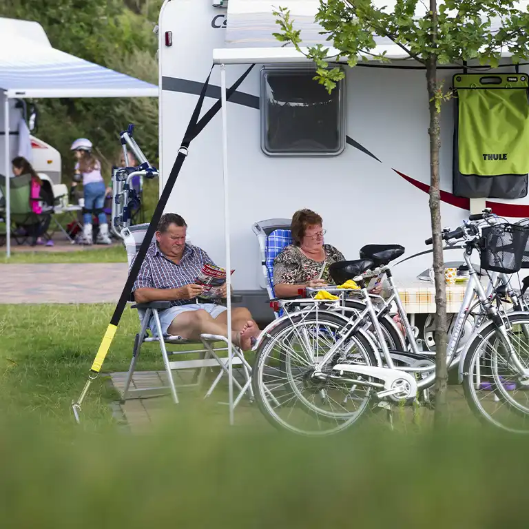 Älteres Paar sitzt entspannt vor einem Wohnwagen unter einem Sonnendach und liest Zeitschriften. Zwei Fahrräder stehen im Vordergrund, im Hintergrund sind weitere Camper und spielende Kinder zu sehen – Campingurlaub mit Komfort und Freizeitgestaltung für Senioren.