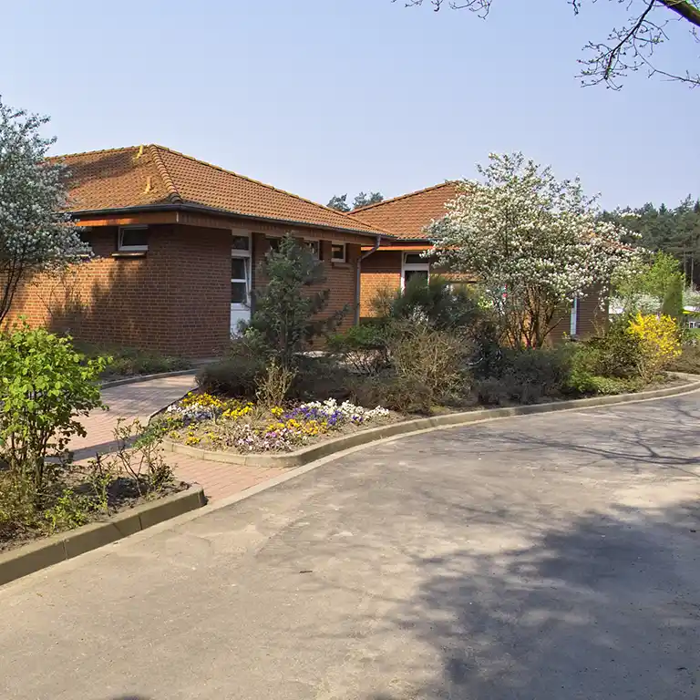 Mehr erfahren Das Waschhaus 10 im Südsee-Camp, ein Backsteingebäude mit rotem Ziegeldach, umgeben von gepflegten Blumenbeeten und blühenden Bäumen – eingebettet in eine grüne, waldnahe Umgebung.