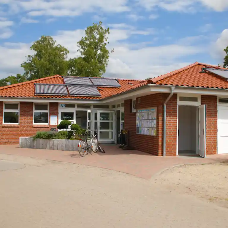 Mehr erfahren Das Waschhaus 2 im Südsee-Camp mit rotem Ziegeldach, Solarpanelen und einem begrünten Eingangsbereich mit Fahrradstellplatz – ein gepflegtes Sanitärgebäude bei sonnigem Wetter.