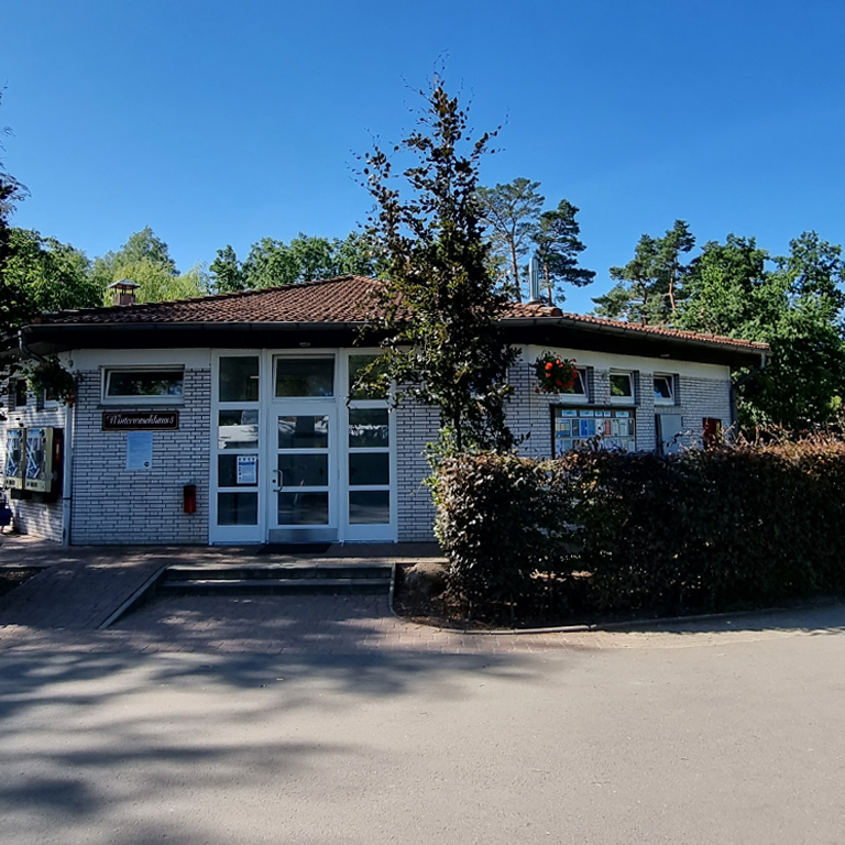 Mehr erfahren Das Waschhaus 5 im Südsee-Camp mit hellen Klinkersteinen, einem braunen Ziegeldach und blühenden Blumen am Fenster, eingerahmt von Bäumen und gepflegten Hecken.