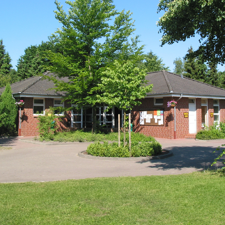 Mehr erfahren Waschhaus 6 im Südsee-Camp, ein rotes Klinkergebäude mit dunklem Walmdach, gepflegtem Vorgarten und Informationstafeln, umgeben von Bäumen und Grünflächen bei strahlendem Sommerwetter.