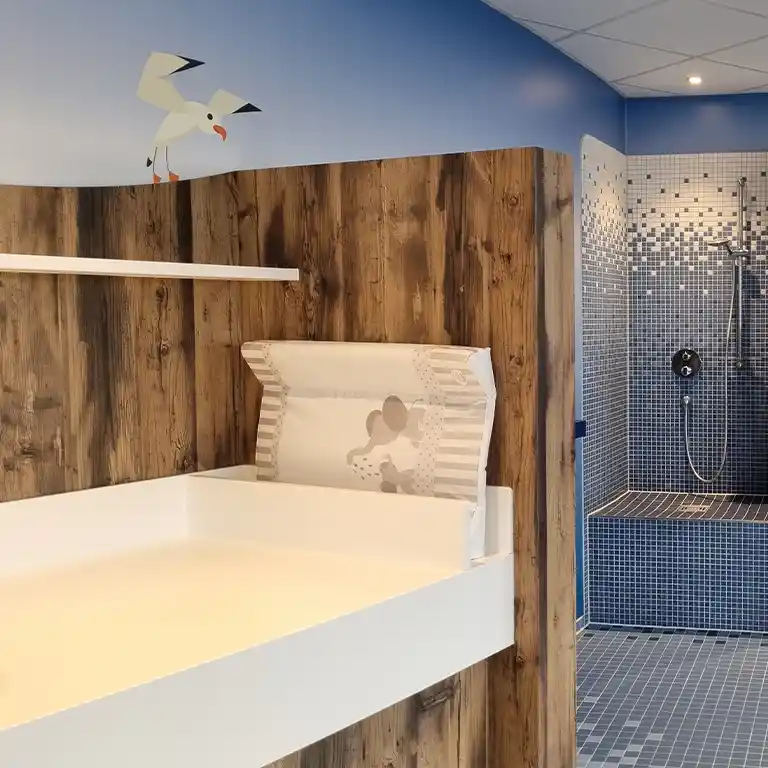 Wickeltisch im Kinderwaschraum des Waschhauses 4 im Südsee-Camp, mit weicher Auflage, Holzverkleidung und einer gefliesten Dusche im Hintergrund. Die Wand ist in Blau gestaltet mit einer Möwen-Illustration.