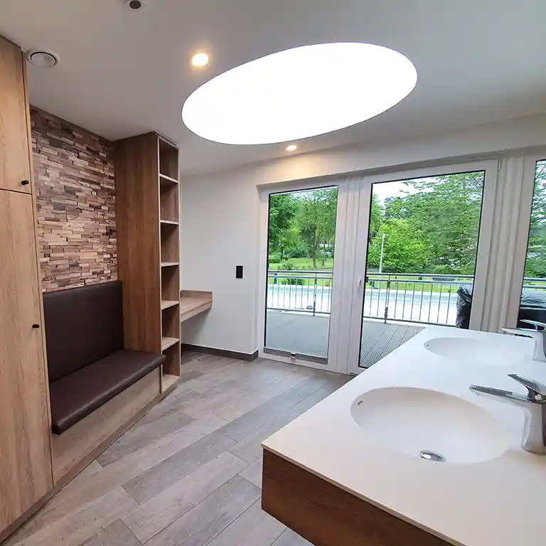 Modernes Privatbad im Waschhaus 4 im Südsee-Camp mit zwei Waschbecken, einer Sitzbank mit Holzverkleidung, Regalfächern, einem Schminktisch sowie einem großen runden Deckenfenster, das Tageslicht hereinlässt. Durch die Glastüren Blick auf die Terrasse und die Grünanlage.