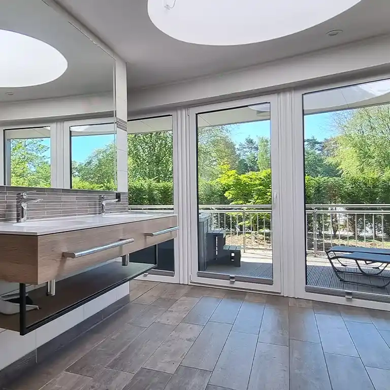 Privatbad im Südsee-Camp mit moderner Doppelwaschbeckenanlage, großen Fenstern und Blick auf eine möblierte Terrasse im Grünen.