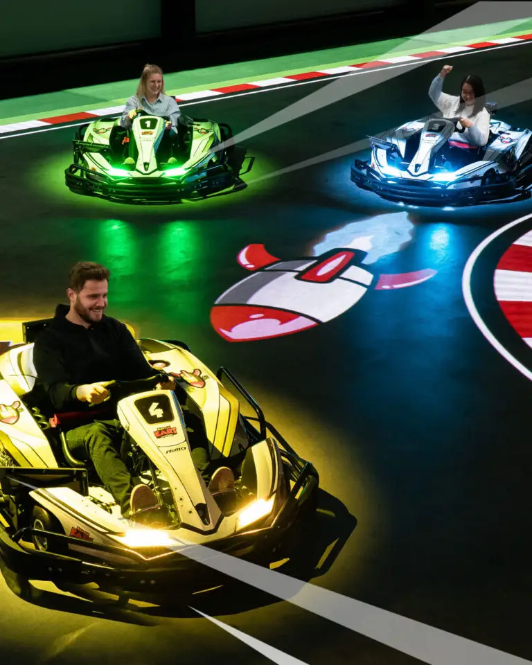 zur augmented Reality Kartbahn Mehrere Personen fahren in leuchtenden E-Karts durch die interaktive BattleKart-Arena in Bispingen. Die Strecke ist farbig beleuchtet, mit digitalen Projektionen wie einem Raketen-Power-up, die das Fahrerlebnis spielerisch erweitern. Die Fahrer*innen wirken begeistert und genießen das actionreiche Ausflugsziel – ideal für Familien und Gäste des Südsee-Camps, die Spaß und Abenteuer suchen.