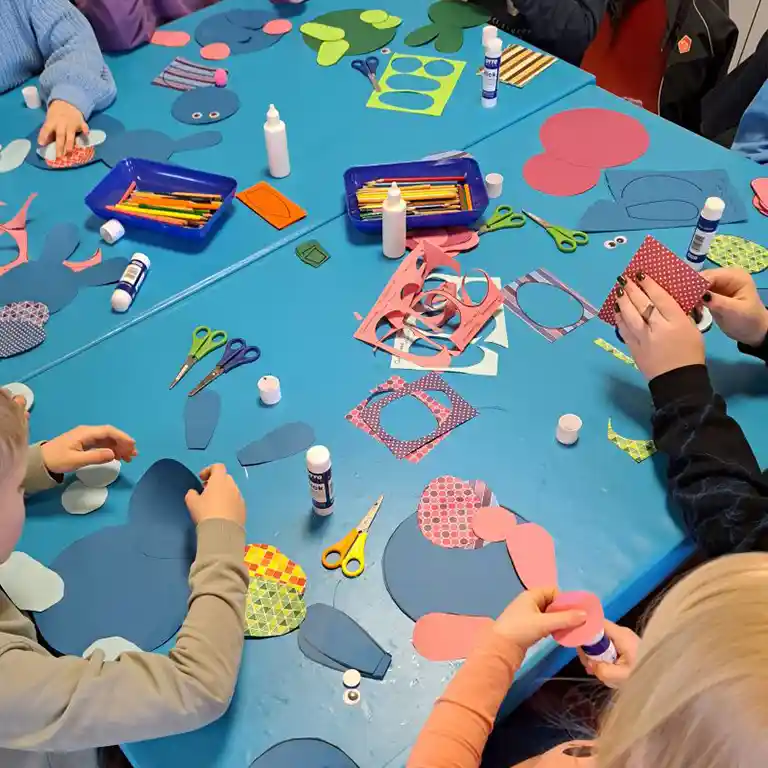 zum Animationsprogramm Kinder sitzen an einem großen, blauen Basteltisch im Südsee-Camp und schneiden bunte Papierformen aus. Auf dem Tisch liegen Scheren, Kleber, Stifte und farbiges Papier in vielen Mustern. Die Kinder gestalten kreative Figuren und genießen eine gemeinsame Bastelaktivität – ein familienfreundliches Angebot für kreative Urlaubsmomente.