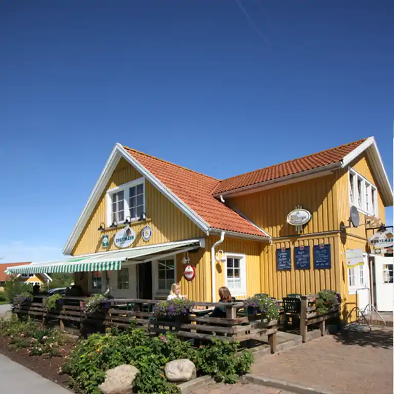 Skandinavisch gestaltetes Restaurant Bootsmann im Feriendorf Sommarby im Südsee-Camp mit gelber Holzfassade, rotem Ziegeldach und Außenterrasse mit Blumenkästen.