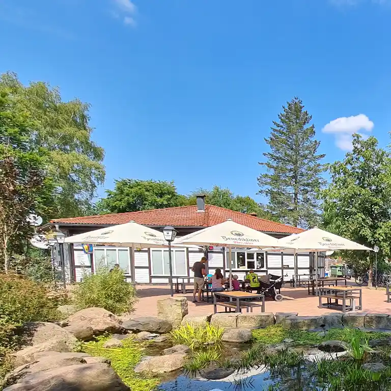 Außenbereich des Imbiss Campers Inn im Südsee-Camp mit schattigen Sitzplätzen unter Sonnenschirmen und einem Teich im Vordergrund bei blauem Himmel.