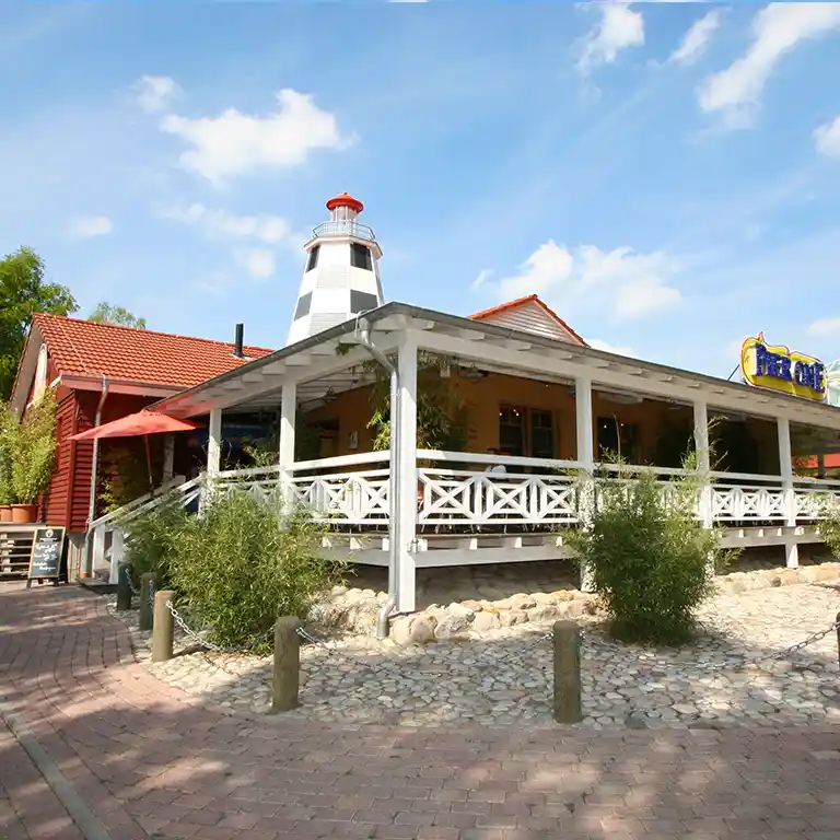 Restaurant Pier One im Südsee-Camp mit großer Veranda, maritimem Leuchtturm und weißer Holzfassade unter blauem Himmel.