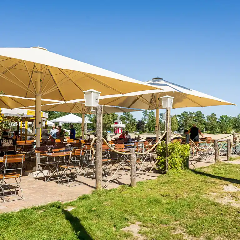 Strandbar am See im Südsee-Camp mit Holzstühlen unter großen Sonnenschirmen, direkt am Sandstrand mit Blick auf das Wasser und roten Leuchtturm.