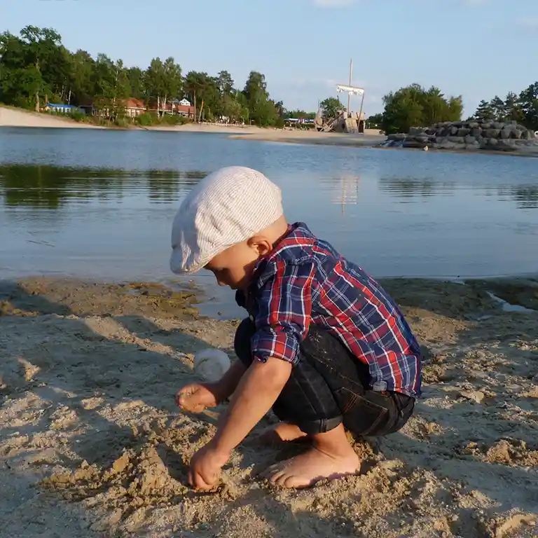 Kind am Strand spielt mit Sand, im Hintergrund ein Badesee