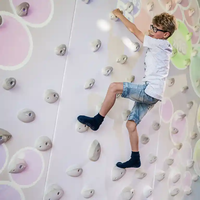 Ein Junge klettert an der virtuellen Kletterwand im Südsee-Camp. Mit 3D-Effekten und interaktiven Griffen bietet die Wand sportlichen Spaß und Abenteuer für Kinder im Familienurlaub.