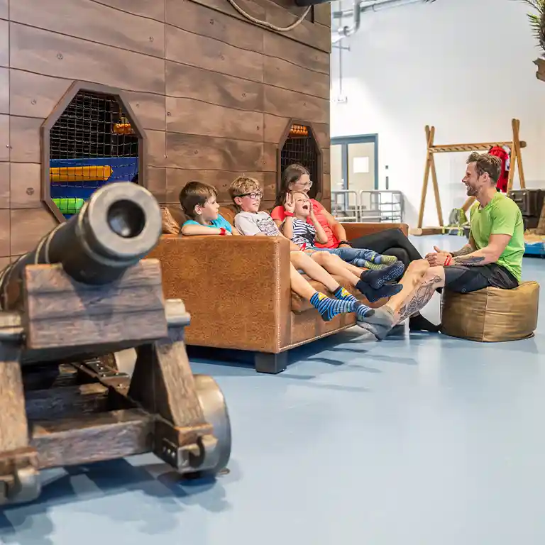Kinder sitzen entspannt auf einem Sofa im Indoorspielplatz „Piratennest“ im Südsee-Camp, während zwei Erwachsene mit ihnen lachen und sprechen. Die Piratenkulisse mit Kanone sorgt für Abenteuerflair – ein Ort für Spiel und Erholung im Familienurlaub.