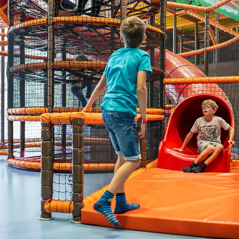 Kinder toben im Indoorspielplatz „Piratennest“ im Südsee-Camp. Ein Junge rutscht lachend aus einer roten Röhre, während ein anderer am Ausgang wartet – pure Action und Spielspaß für Familien mit Kindern.