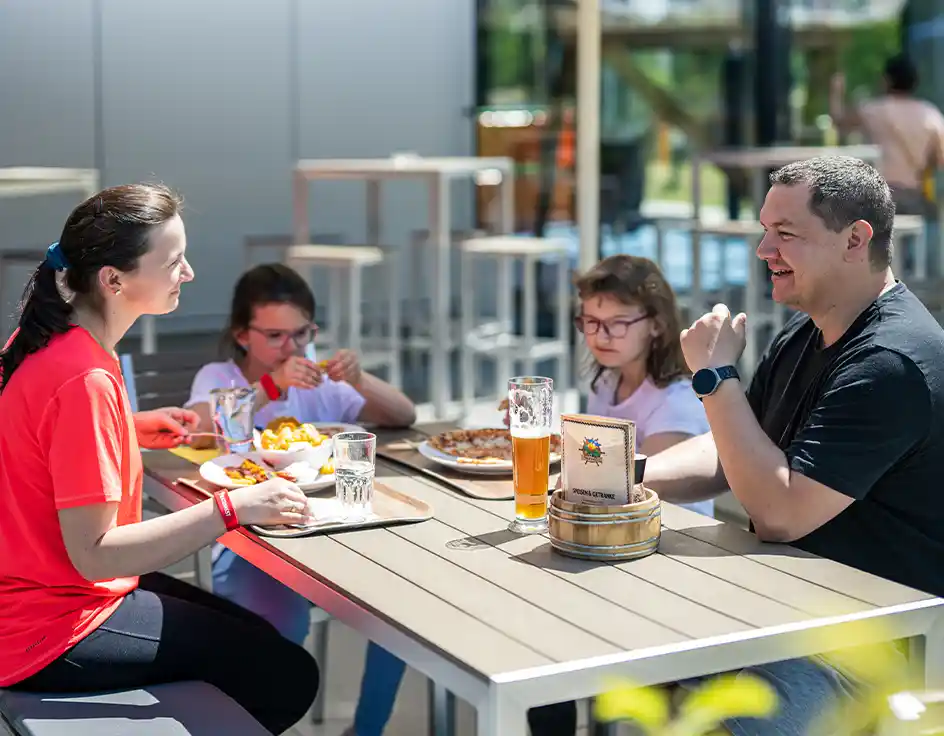 Essen & Trinken: Restaurants, Bars & Imbisse im Südsee-Camp Wietzendorf Familie sitzt bei sonnigem Wetter auf der Terrasse des Piratennests im Südsee-Camp, isst gemeinsam Pommes und Pizza an einem modernen Holztisch.