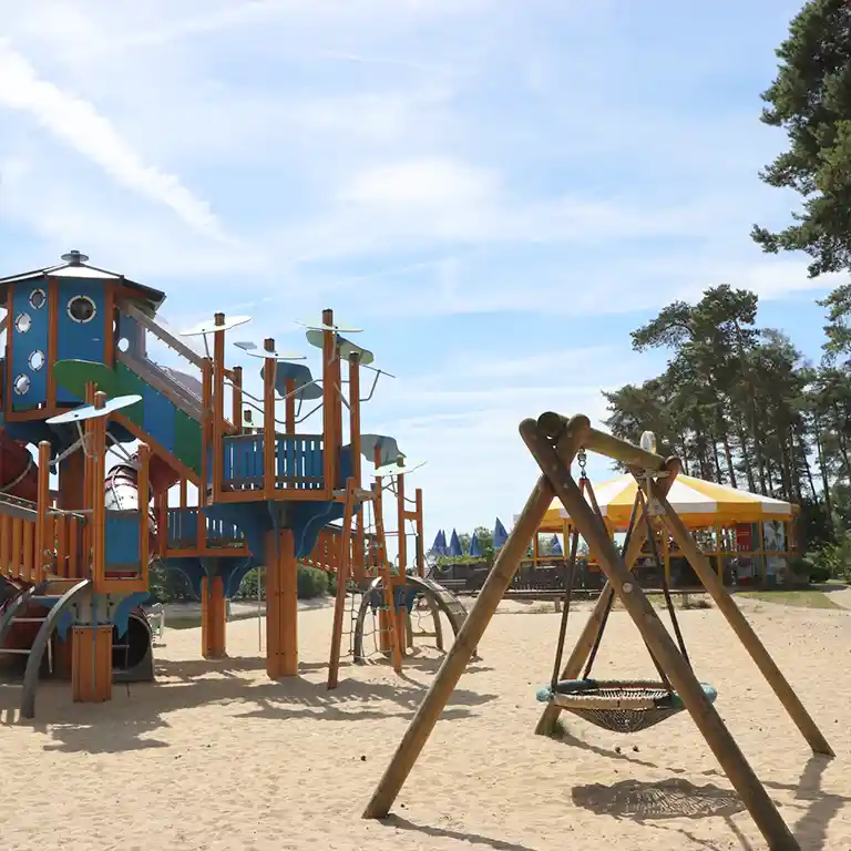 Mehr erfahren Großer Abenteuerspielplatz mit Rutschenturm, Kletterelementen und Nestschaukel auf Sandfläche im Südsee-Camp – ideal für Familien mit Kindern.
