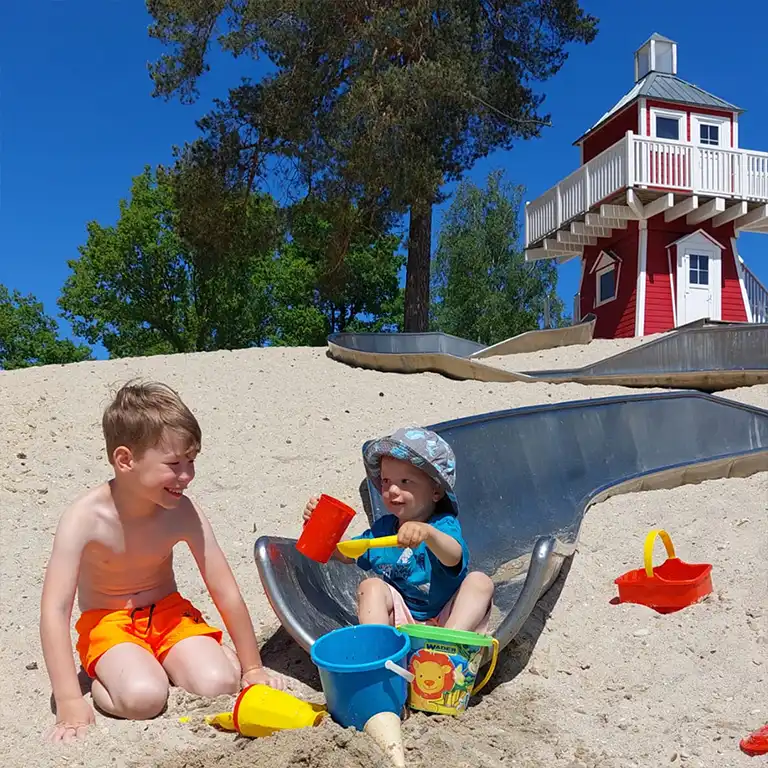 Zwei Kinder spielen fröhlich im Sand am Leuchtturm-Spielplatz im Südsee-Camp. Während eines entspannten Familientags genießen sie das schöne Wetter, rutschen und bauen mit bunten Sandspielzeugen – ideal für kleine Entdecker.
