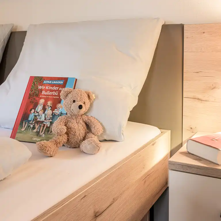 Elternbett in einem Schwedenhaus im Südsee-Camp – auf dem Bett liegen ein hellbrauner Teddybär und das Kinderbuch „Wir Kinder aus Bullerbü“ von Astrid Lindgren, gemütliche und familienfreundliche Atmosphäre.