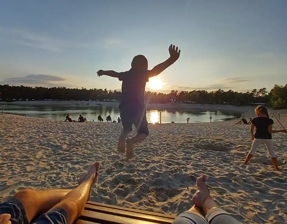 You've booked Child jumping at the sandy beach in the evening sun in front of the lake at Südsee-Camp; in the foreground, relaxed feet of two people on a bench