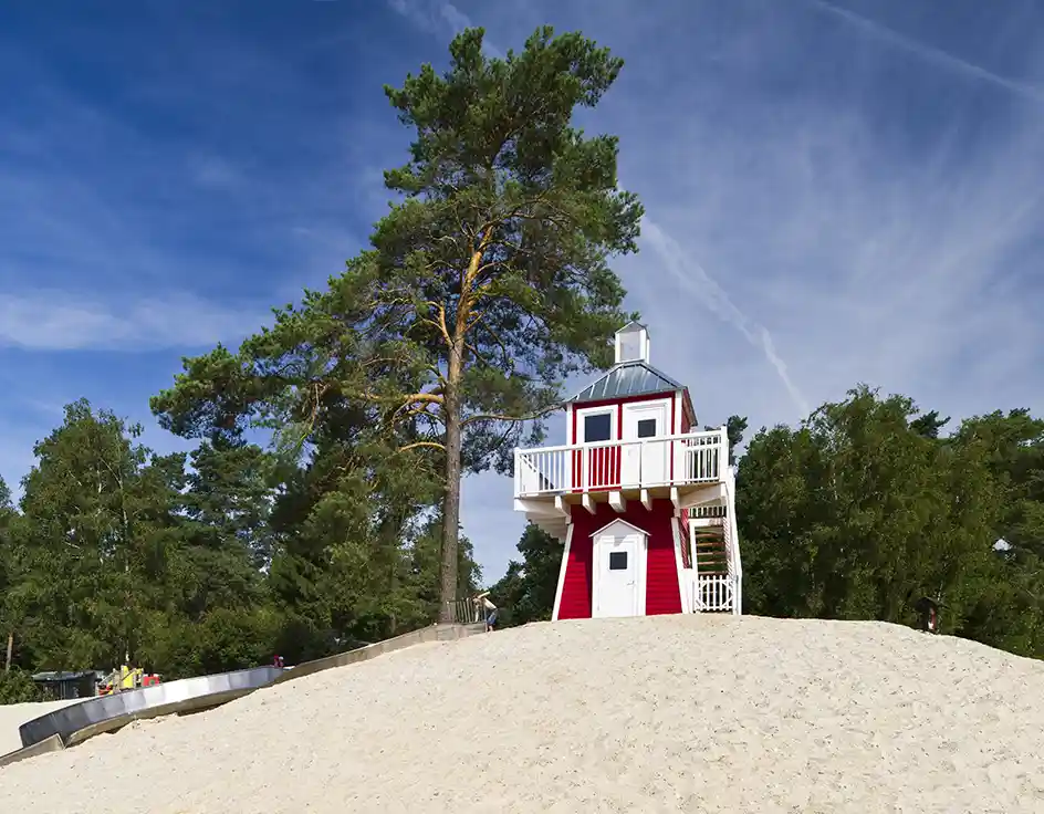 Prices 2025 & 2026: Cottages, chalets, rental caravans | Südsee-Camp Red lighthouse with white balcony at the beach of Südsee-Camp, next to a long slide and surrounded by pine trees under a blue sky