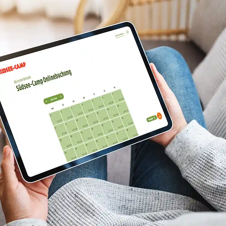 A person holds a tablet and uses the Südsee-Camp online booking system. The screen shows the digital site map for holiday planning – convenient booking from home.