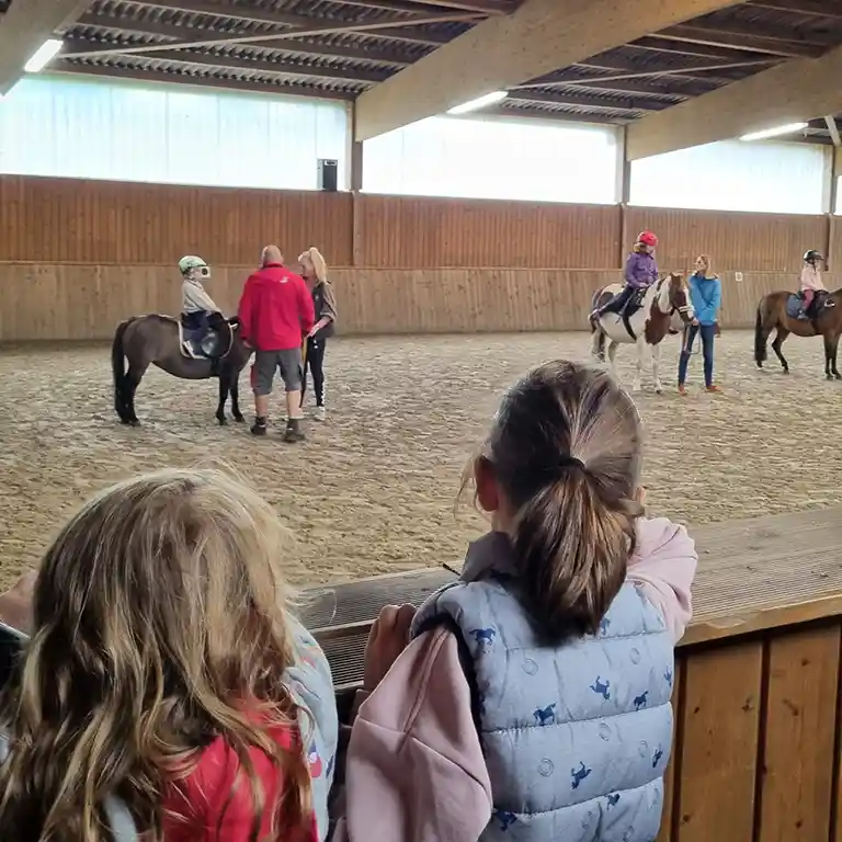 Kinder schauen vom Rand der Reithalle im Südsee-Camp zu, wie andere Kinder auf Ponys reiten. Betreuer geben Hilfestellung und sorgen für Sicherheit – ein spannendes Ferienerlebnis für Familien.