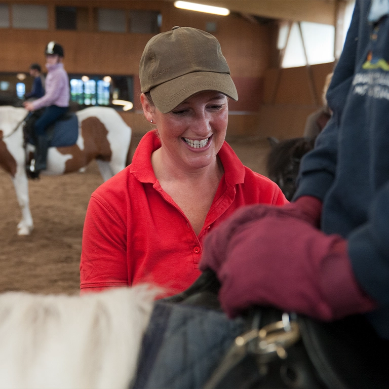 Eine Reitlehrerin im roten Shirt lächelt während des Unterrichts in der Reithalle des Südsee-Camps. Im Hintergrund reiten Kinder auf Ponys – ein fröhliches Erlebnis für Familien im Urlaub.