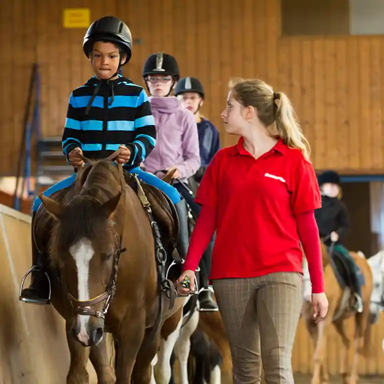 Kinder nehmen an einer Reitstunde im Südsee-Camp teil. Ein Junge reitet auf einem Pferd und wird dabei von einer Betreuerin in rotem Shirt geführt. Weitere Kinder folgen auf Ponys in der Reithalle – ein spannendes Ferienerlebnis für Familien.