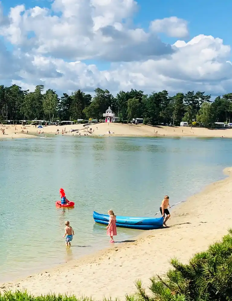 auf zum Strand! Kinder am flachen Sandstrand des Badesees mit Ruderboot und Wald im Hintergrund