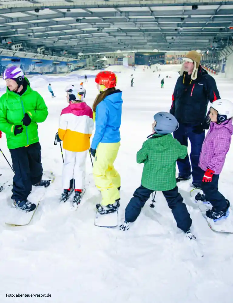 to the ski hall Family in colorful winter gear snowboarding and skiing inside SnowWorld Bispingen – an indoor ski paradise for kids and adults in all weather conditions.