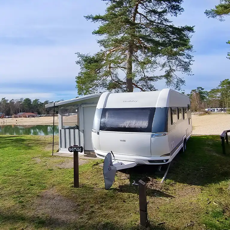 zu den Mietwohnwagen Weißer Mietwohnwagen mit Vorzelt auf Stellplatz am See, im Hintergrund Sandstrand und Bäume