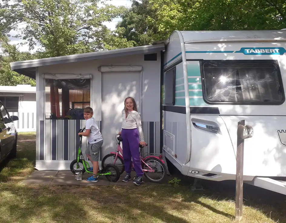 Holidays in a rental caravan at Südsee-Camp Two children stand with their scooters and bikes in front of a rental caravan with a fixed awning at Südsee-Camp. The caravan is labeled “Dachs.” A parked car is visible on the left. The scene takes place under trees in daylight.