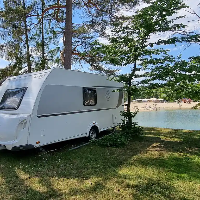 White Knaus Sport caravan parked in the shade directly by the lake at Südsee-Camp. In the background, there’s a sandy beach with sun umbrellas and people enjoying the water under a blue sky with a few clouds.