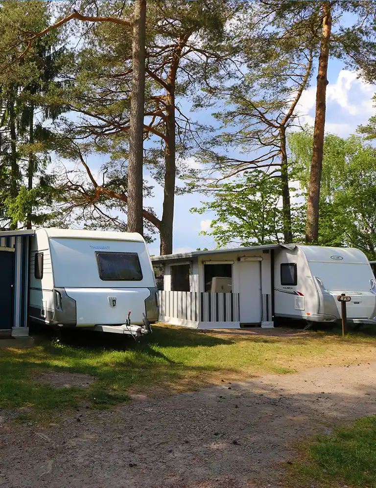 Learn more Two white rental caravans with fixed awnings at Südsee-Camp, set under tall pine trees. A wooden sign labeled “Bär” stands between the caravans. The ground is a mix of grass and sand, with trees and a slightly cloudy sky in the background.