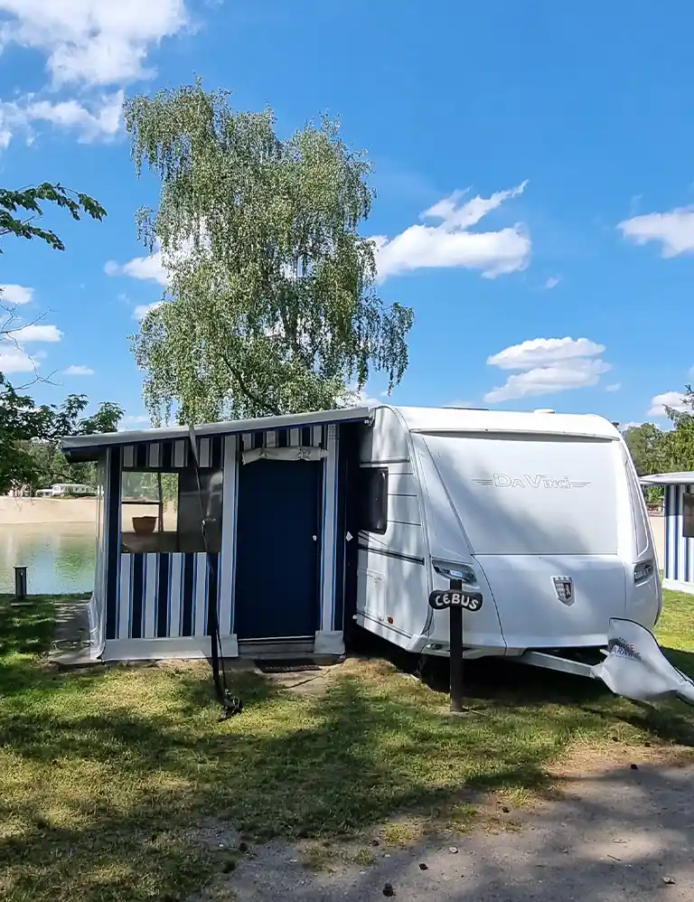 Learn more White rental caravan with a fixed blue-and-white striped awning at Südsee-Camp. In the background, there is a small lake with a sandy beach. A sign in front of the caravan reads “Cebus.” A large tree provides partial shade.