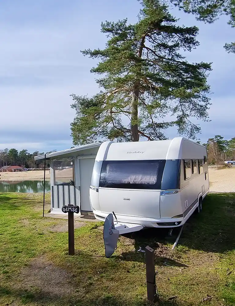 Learn more White rental caravan with a fixed awning at Südsee-Camp, located directly by the water. In the foreground, a wooden sign labeled “GEMSE” and a satellite dish. A sandy beach and trees are visible in the background.